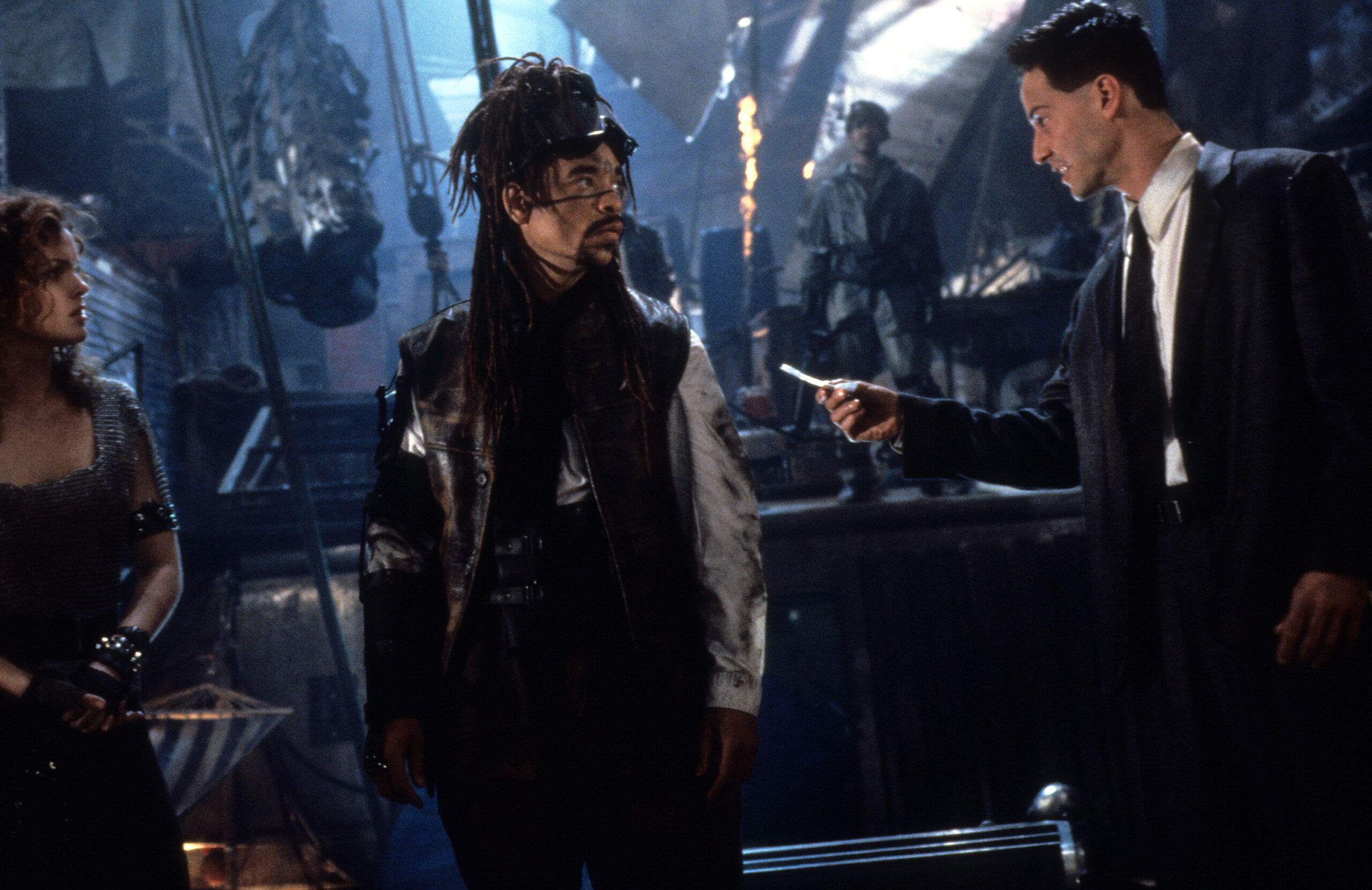 Three people stand in a dark, smoky club as a man in a leather vest and dreads looks toward a man in a suit who offers him a blade or knife.