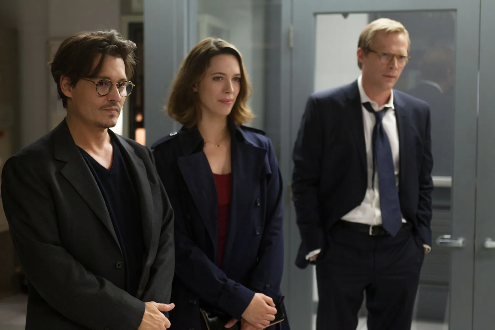 Three professionals stand in a modern office corridor, wearing dark business attire and looking ahead pensively.