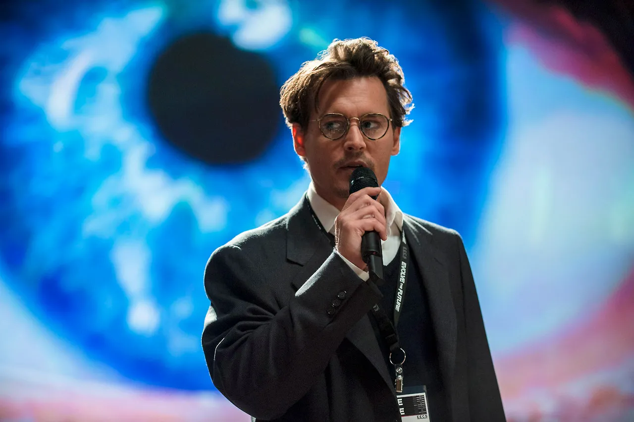 Man in a dark suit speaks into a handheld microphone on a stage with a blue abstract backdrop.