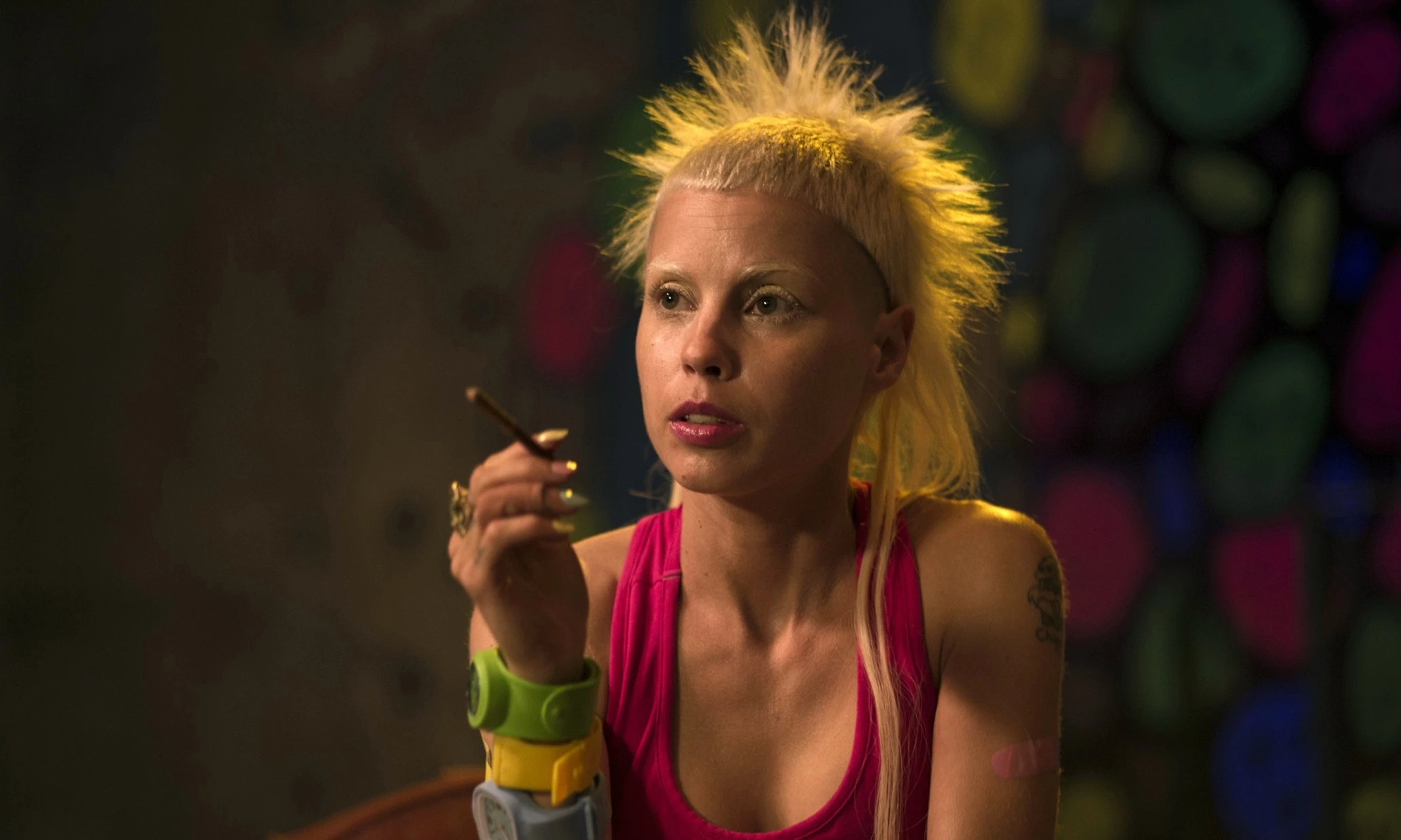 Woman with spiky blonde hair in a pink top lighting a cigarette in a dim setting.
