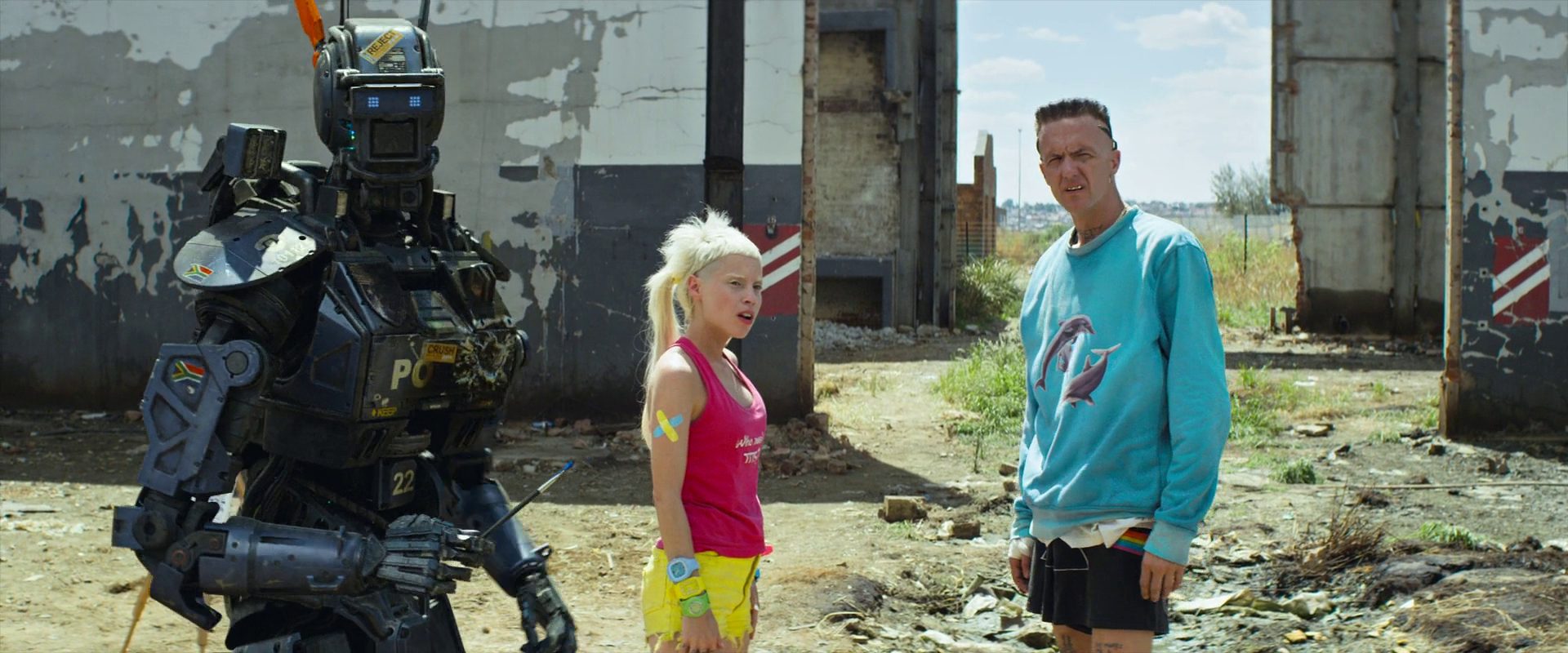A large futuristic robot stands with a blonde woman in a pink top and yellow shorts and a man in a teal sweater at a ruined industrial site.