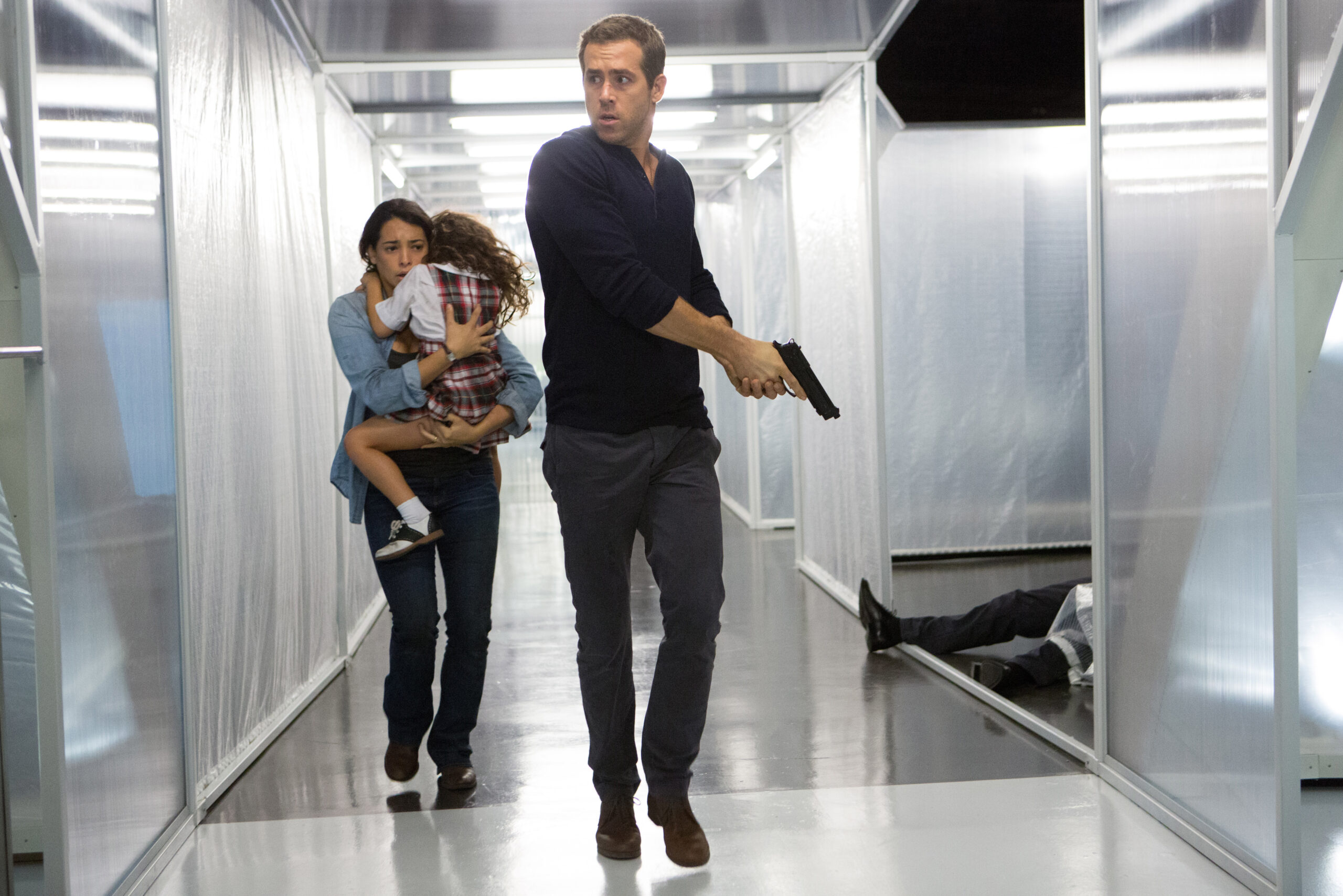 Man in a dark shirt walks toward the camera, holding a handgun, as a distressed woman carries a child behind him; another person lies on the floor in the background in a metallic hallway.