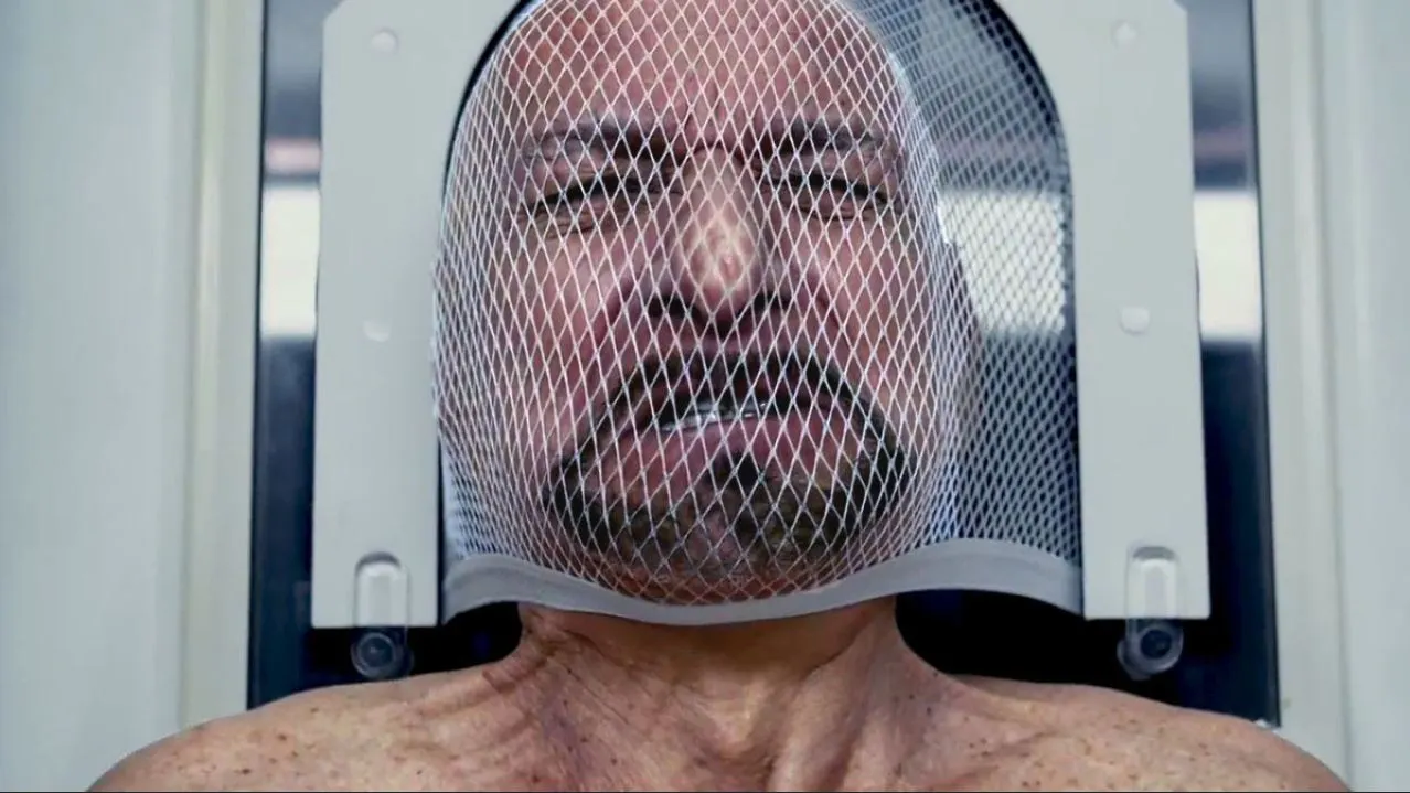 Man wearing a white mesh hood attached to a metal frame, looking distressed.