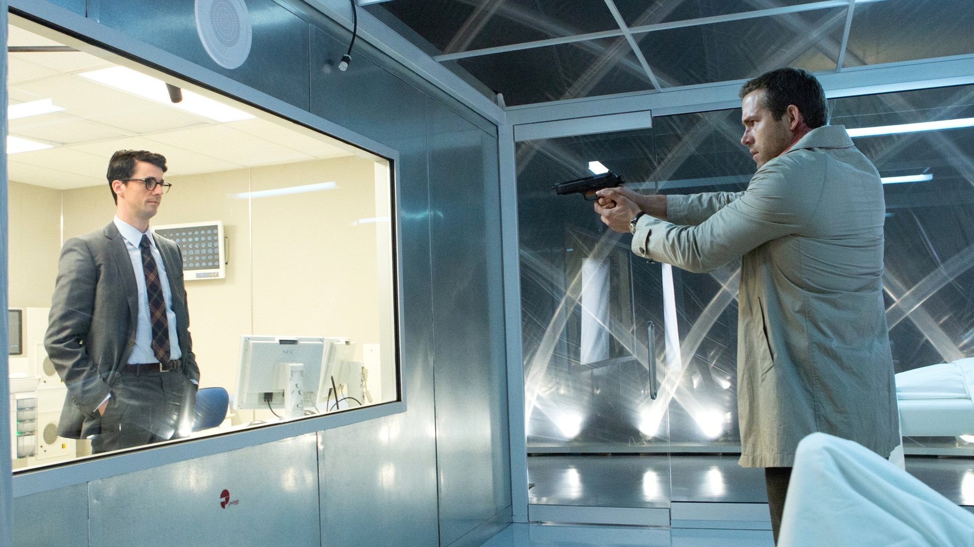 Man in a gray suit stands behind a glass partition while another man in a beige coat aims a pistol at him through the glass.