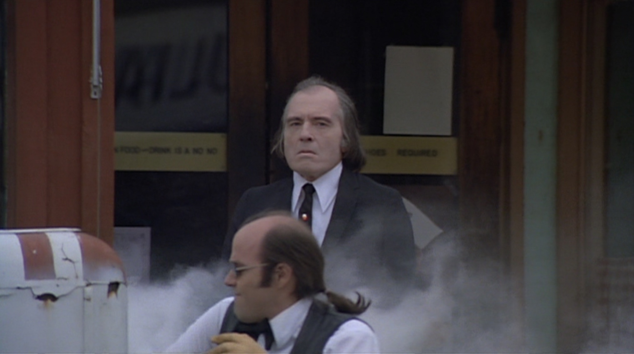 Screenshot | film.org.pl Man in a suit standing behind glass doors in a smoky doorway, looking forward, with a blurred person in front.