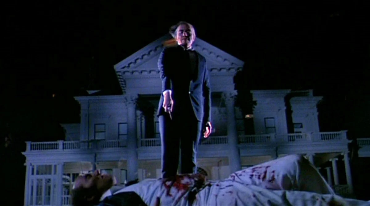 Phantasm-8 | film.org.pl A man in a dark suit stands over a bloodied body on the ground in front of a large white mansion at night.
