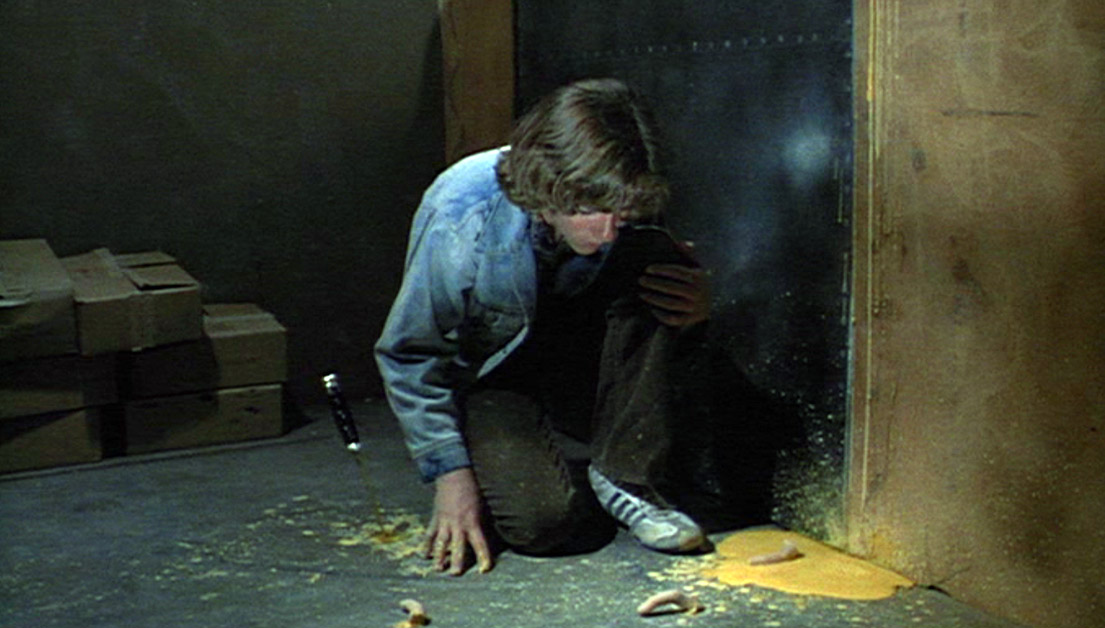 Phantasm-9 | film.org.pl Person kneeling on a dim floor, looking at a phone while a yellow powder shadow lies near a wall or doorway in a cluttered room on the left.