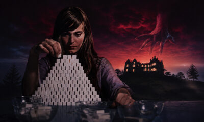 Woman stacks a pyramid of white sugar cubes at a table, with a looming red sky and a giant hand reaching from the clouds toward a distant haunted house.