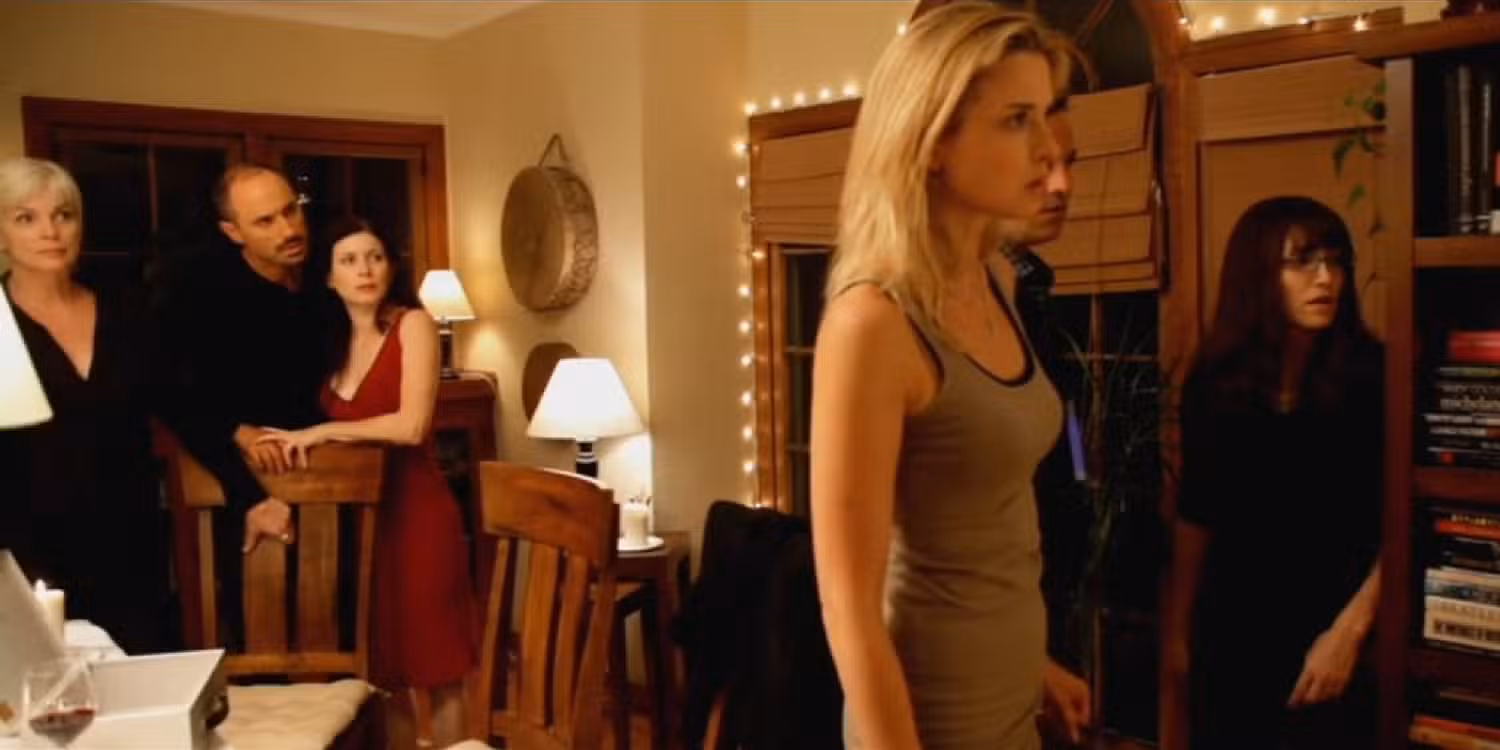 Blonde woman in a beige tank top stands in the foreground as a group of four watches in a warmly lit dining-room setting with string lights.