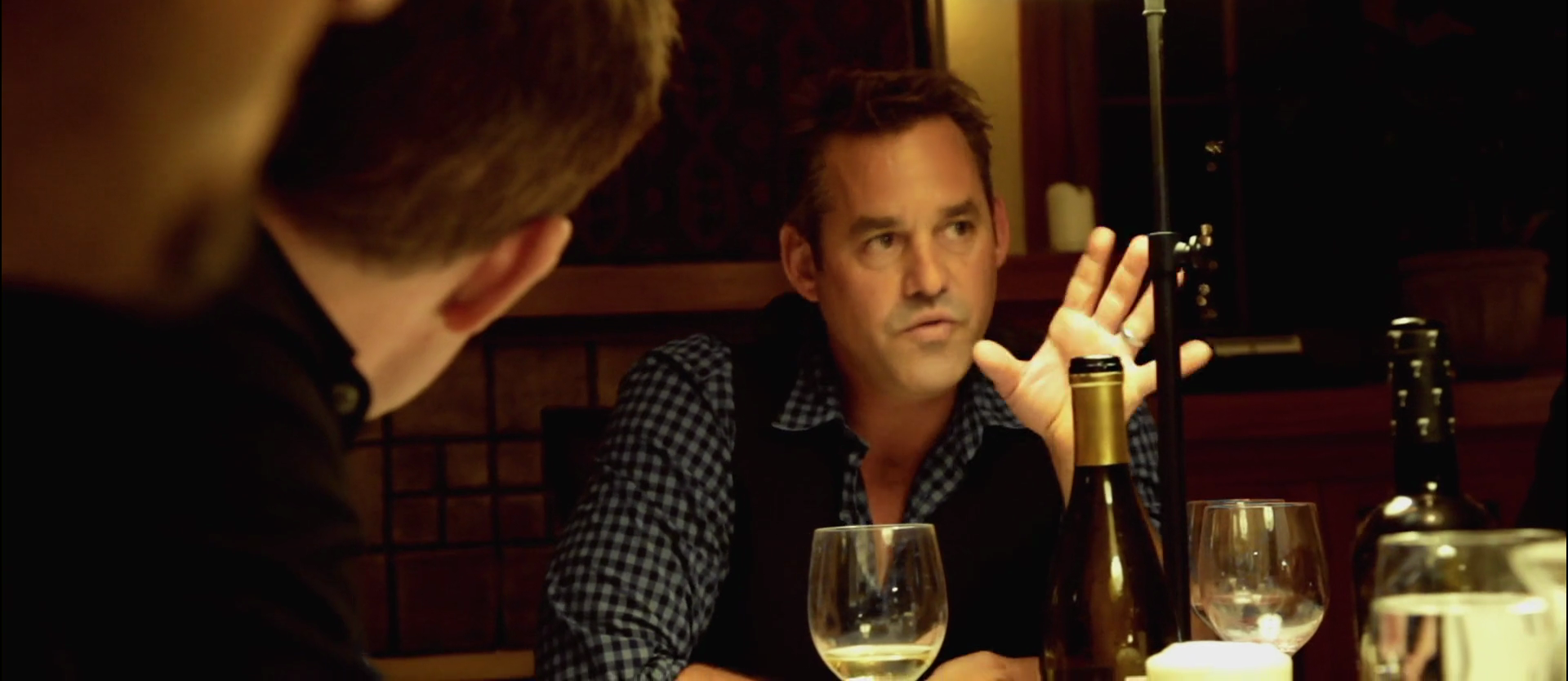 Man in a checkered shirt speaks, gesturing with his hand at a dinner table with wine glasses and bottles nearby