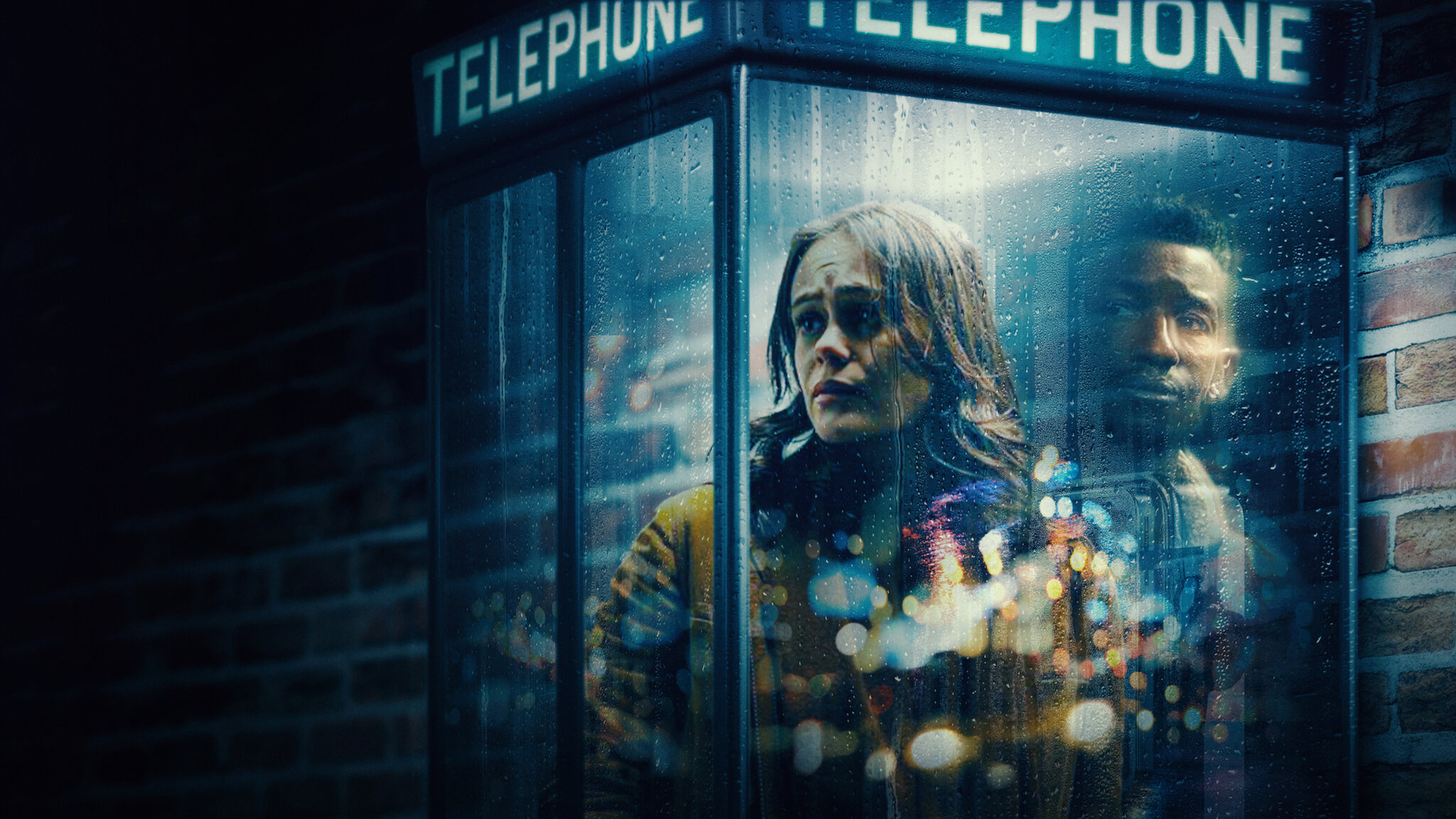 Two people huddle inside a rain-soaked telephone booth at night, looking anxious and tense.