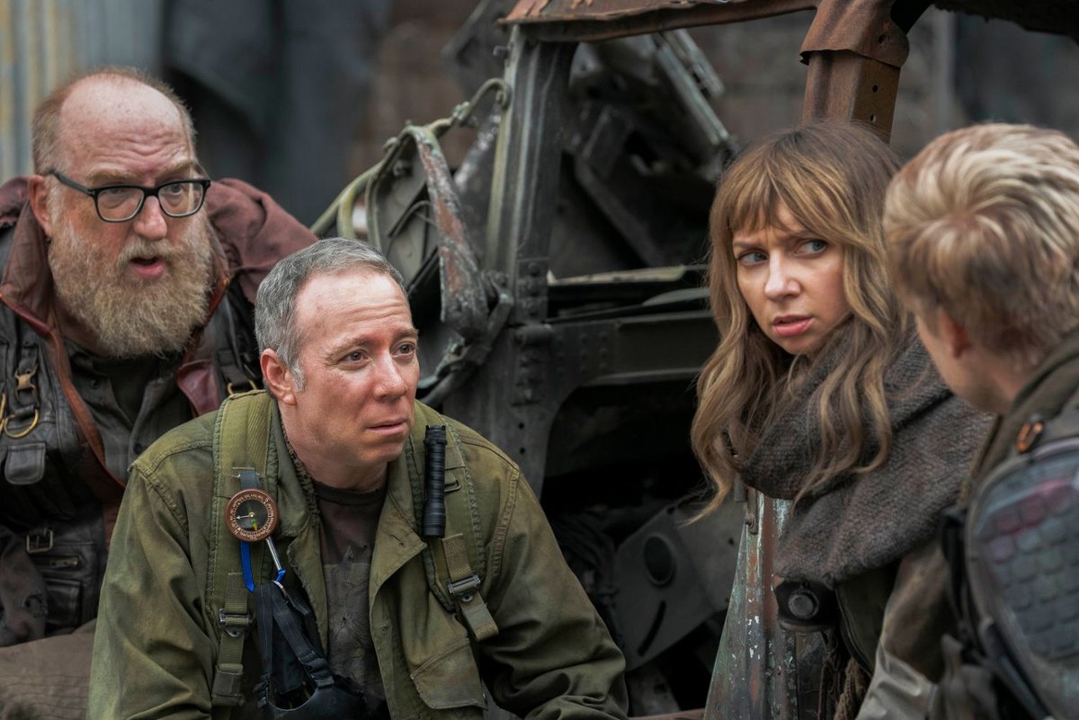 Group of four people in rugged gear crouching beside a wrecked machine, looking tense as they talk.