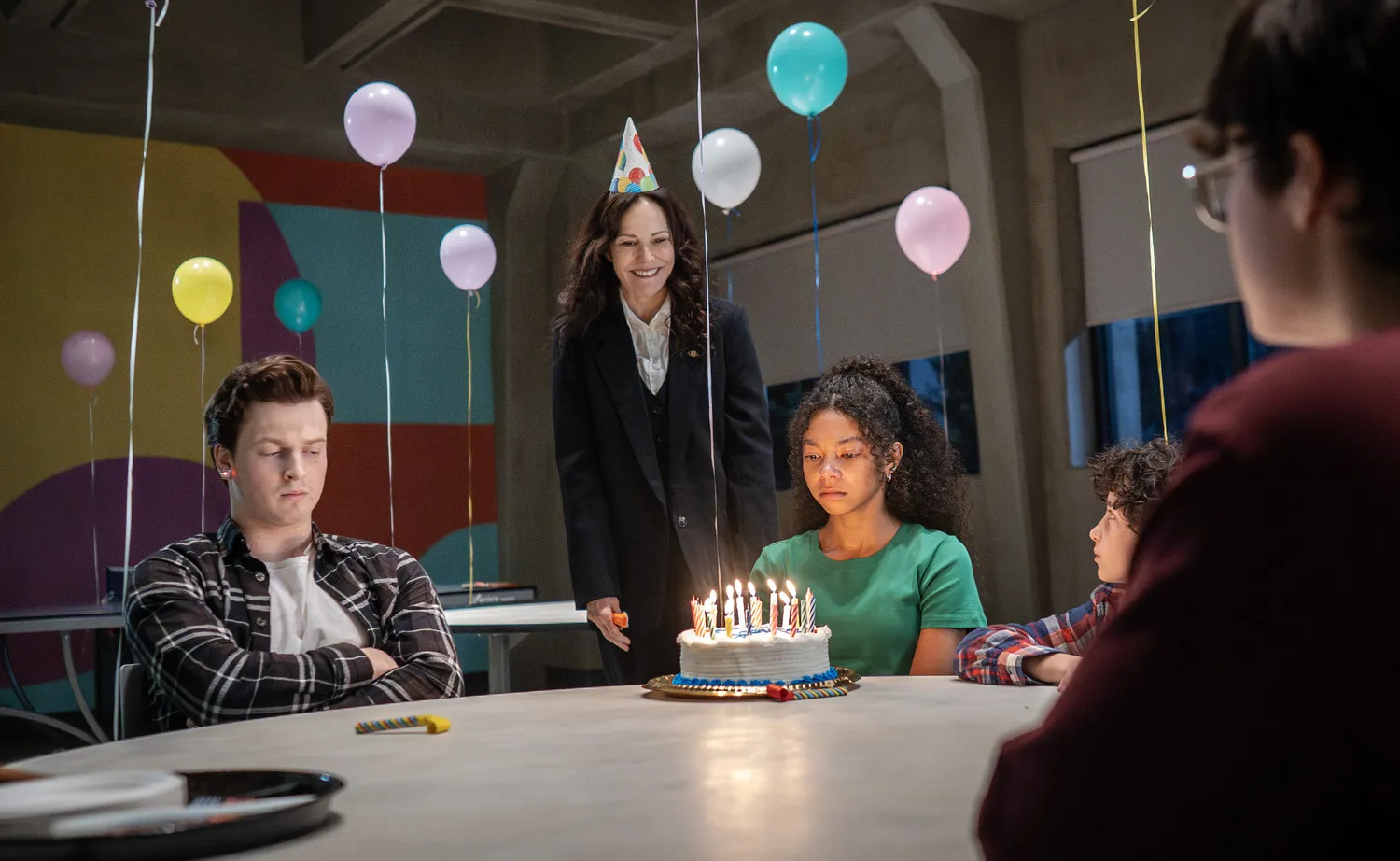 The-Institute-3 | film.org.pl A woman wearing a party hat stands behind a birthday cake with lit candles as friends sit around a table under colorful balloons.