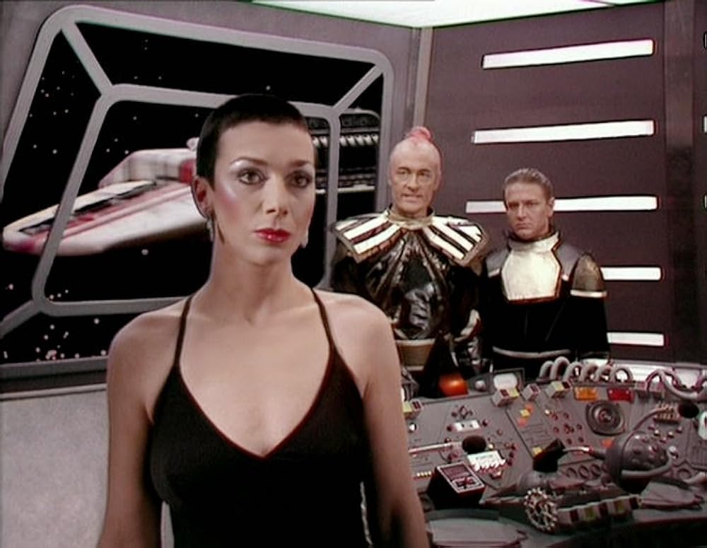 Woman in a black halter dress stands at the front of a sci‑fi spaceship bridge with two armored officers in the background, control panels around them.