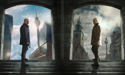 Two men stand on opposite sides of a stone arch doorway, facing each other across a central seam, in a split-city backdrop of modern and historic architecture on a stone staircase.