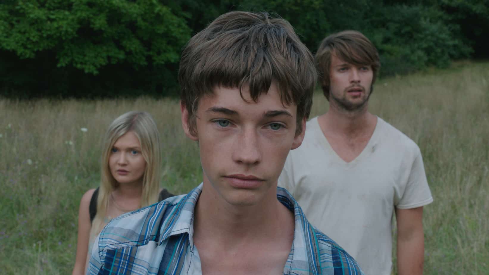 Three young adults stand in a grassy field with trees in the background; the foreground man wears a blue plaid shirt and looks at the camera, with a blonde woman and another man behind him.