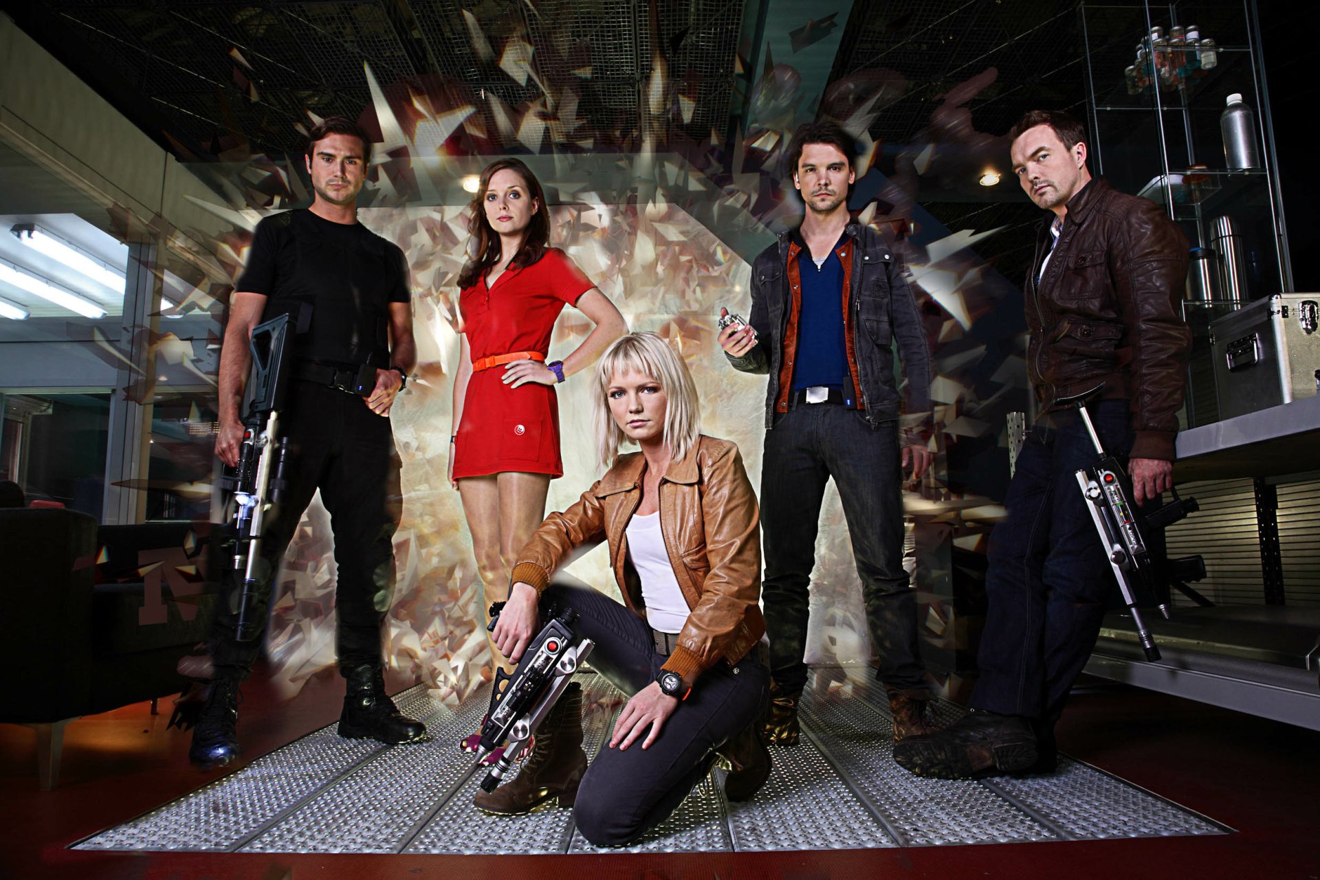 Five people in a futuristic studio pose with prop laser rifles against a shattered, mirrored backdrop.