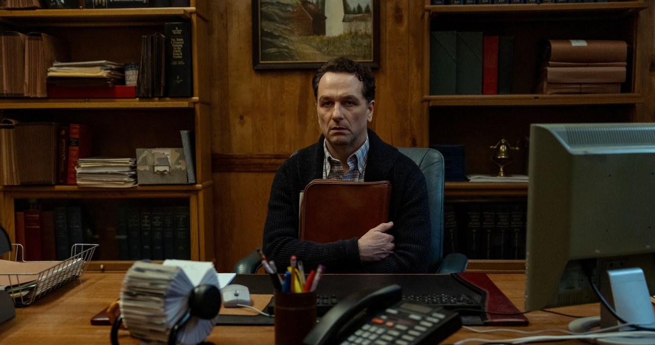Man sits at a large wooden desk in a library-style office, arms wrapped around a brown leather folder, looking serious.