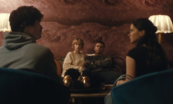Four adults in a cozy, dimly lit living room; two sit on a sofa facing the camera (woman in a cream sweater, man in gray), while two others sit with their backs to the viewer in the foreground.