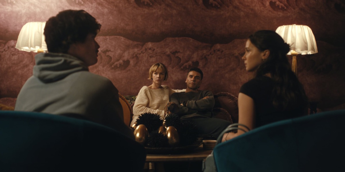 Four adults in a cozy, dimly lit living room; two sit on a sofa facing the camera (woman in a cream sweater, man in gray), while two others sit with their backs to the viewer in the foreground.