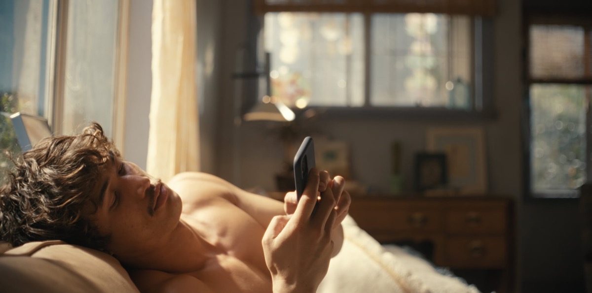 Young man shirtless, lying in bed and scrolling on his smartphone in a sunlit bedroom.