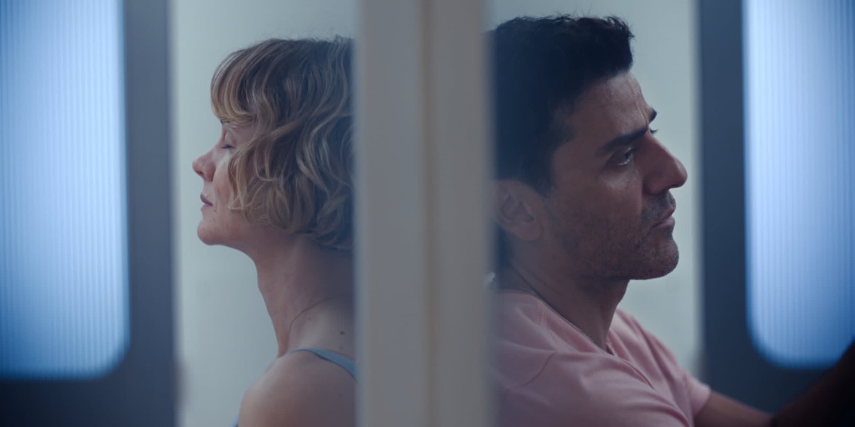 Two adults sit back-to-back separated by a vertical column in a blue-lit corridor, eyes closed and reflective.