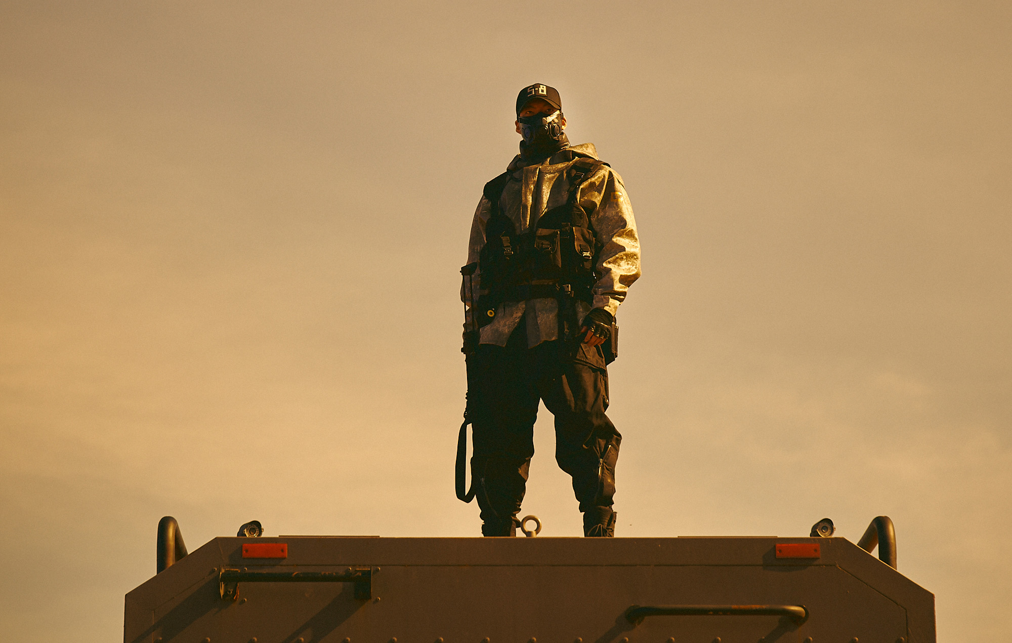 Tactical operator wearing a gas mask and protective gear stands on a vehicle against a warm sunset sky.