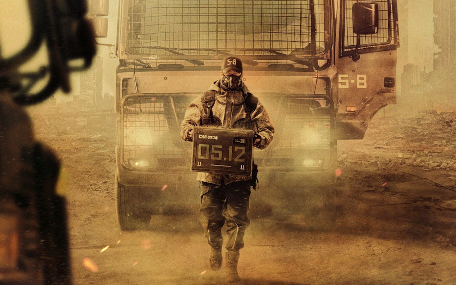 Firefighter in turnout gear and respirator holding a sign reading 05.12 as a dusty fire truck looms behind him.