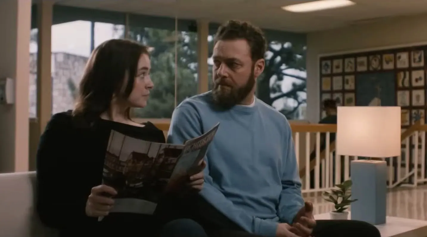 Descendent | film.org.pl A woman and a bearded man sit on a couch in a bright lobby, reading a magazine together.