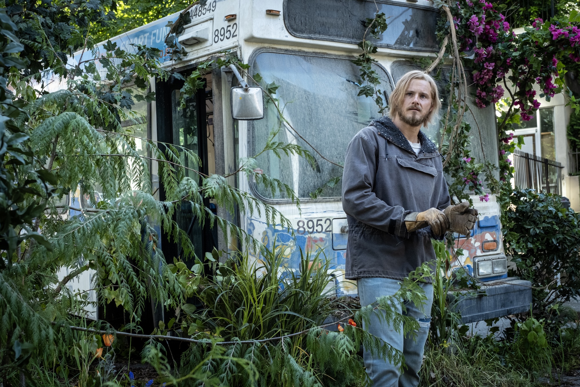 Man in a gray work jacket and gloves stands beside an old, graffiti-tagged bus overgrown with vines and pink flowers in a yard.