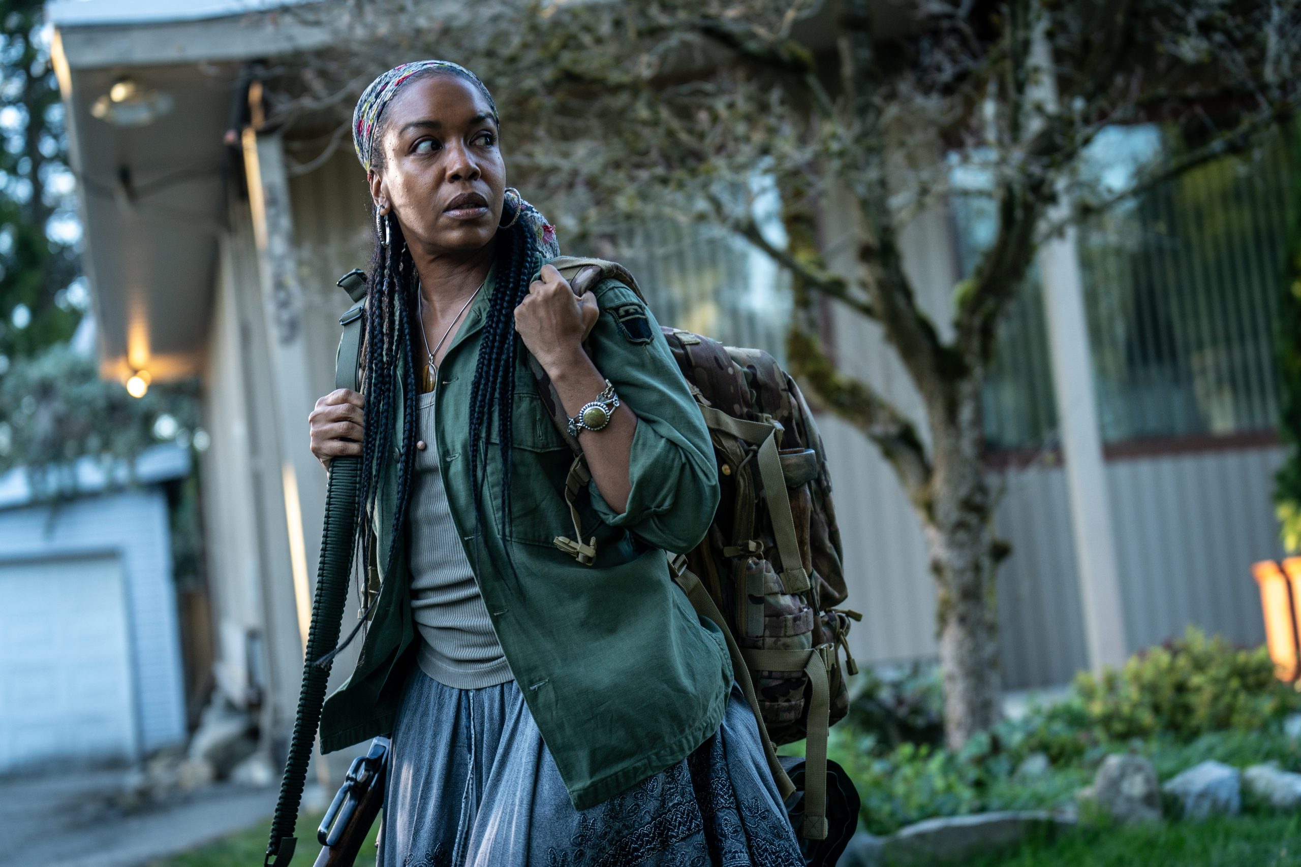 Woman with braided hair and a patterned headscarf wearing a green jacket and backpack, looking to the side on a residential street.