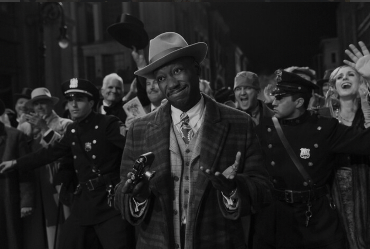 IMG_1469 | film.org.pl Man in a fedora and plaid suit at a festive street parade, surrounded by cheering spectators in period costumes.