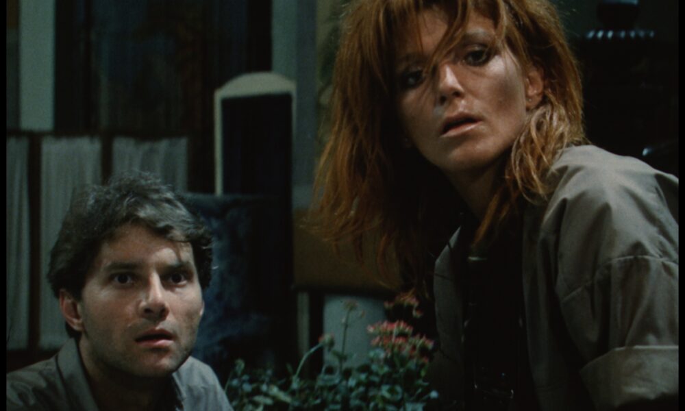 Two adults crouching and looking upward with surprised expressions in a dimly lit room; plants visible in the foreground.