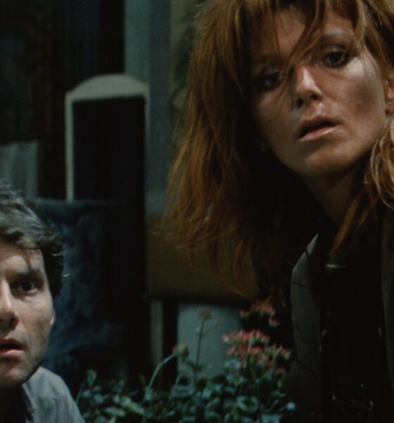Two adults crouching and looking upward with surprised expressions in a dimly lit room; plants visible in the foreground.