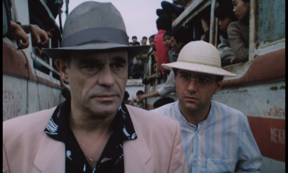 Two men in hats walk past a crowded bus or vehicle, faces serious as onlookers crowd in the background.