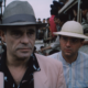 Two men in hats walk past a crowded bus or vehicle, faces serious as onlookers crowd in the background.