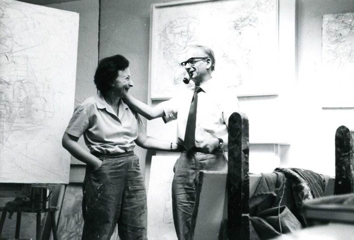 Moi-Themersonowie-3 | film.org.pl Two coworkers in a design studio laugh and chat, with large maps on the wall behind them.