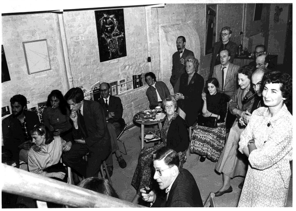 Slajd 1 | film.org.pl Black-and-white photo of a crowded informal gathering in a basement or gallery, with people seated along the wall and several standing, chatting and holding drinks around a small table with pastries and cups. A painting hangs on the wall above the group.