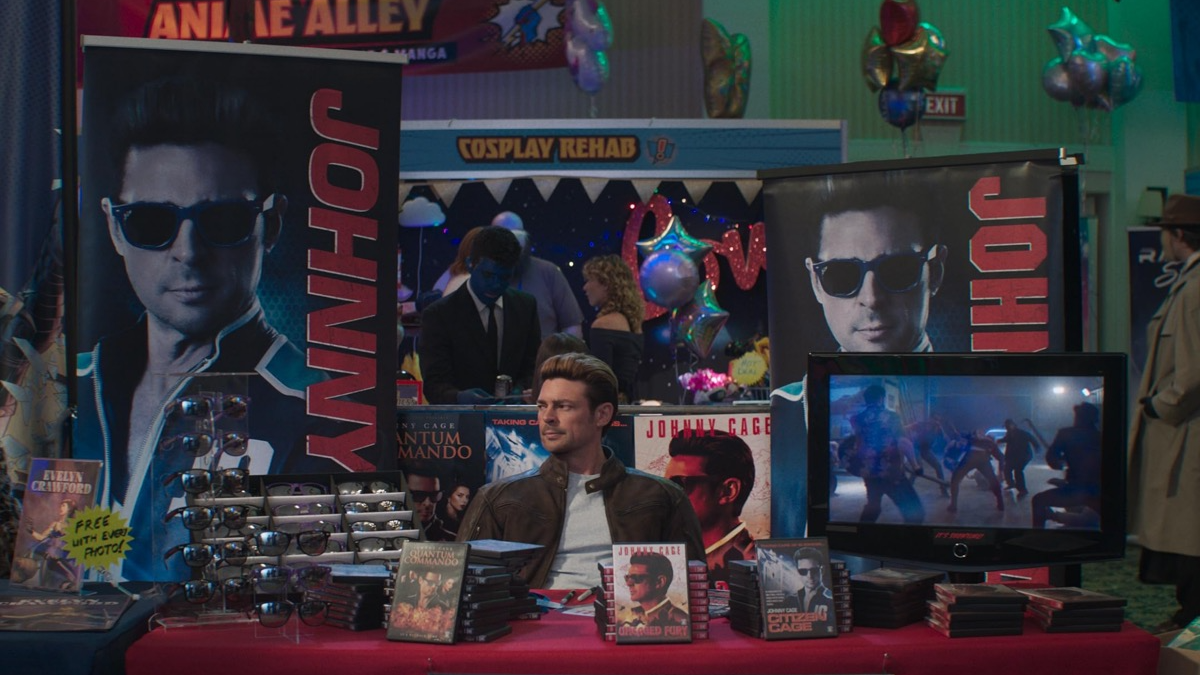 Promotional booth at a convention with large Johnny Cage banners, sunglasses displays, and a man selling DVDs at a merch table.