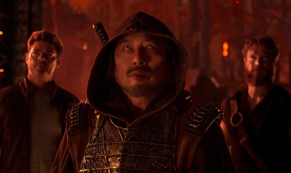 Three hooded warriors in metallic armor stand in a fiery, torch-lit forest; central figure looks upward. 125 chars max approx, but keep concise for screen readers.