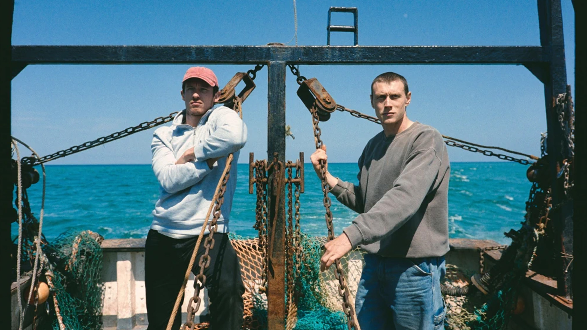 Two men stand on a rusted fishing boat deck, gripping chains with the blue sea behind them.