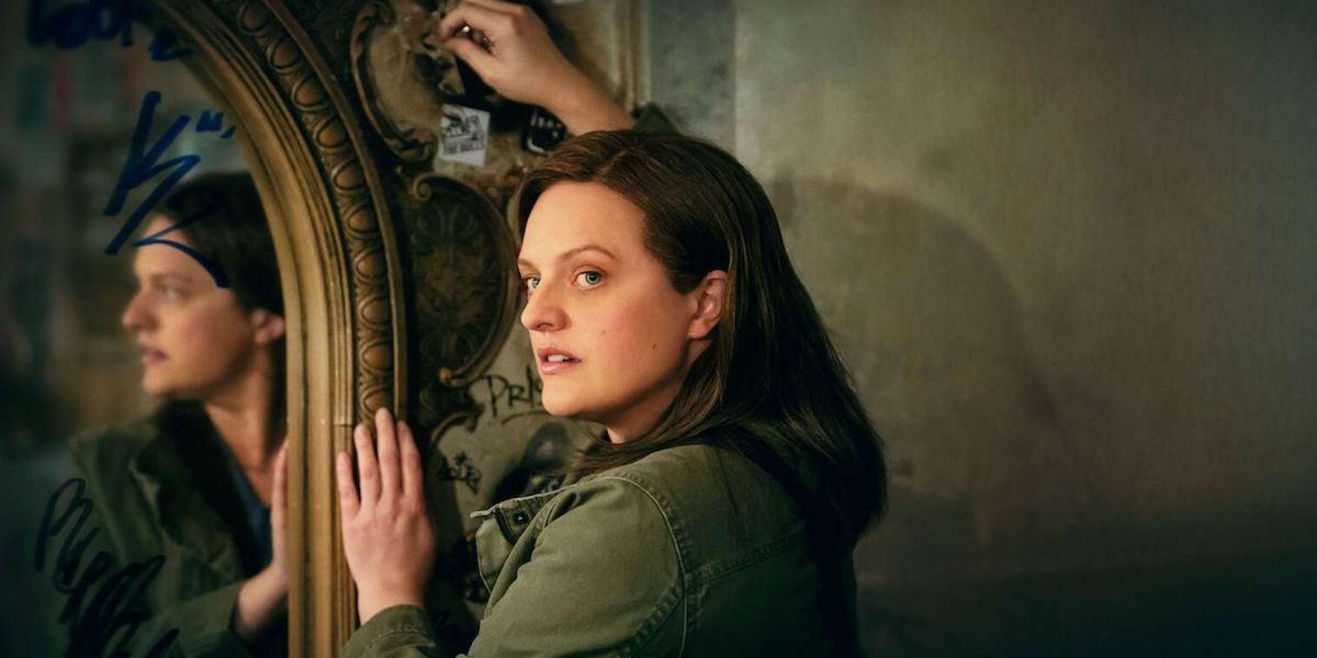 Woman in a green jacket touches a gilded, graffiti-covered mirror and looks at the camera.
