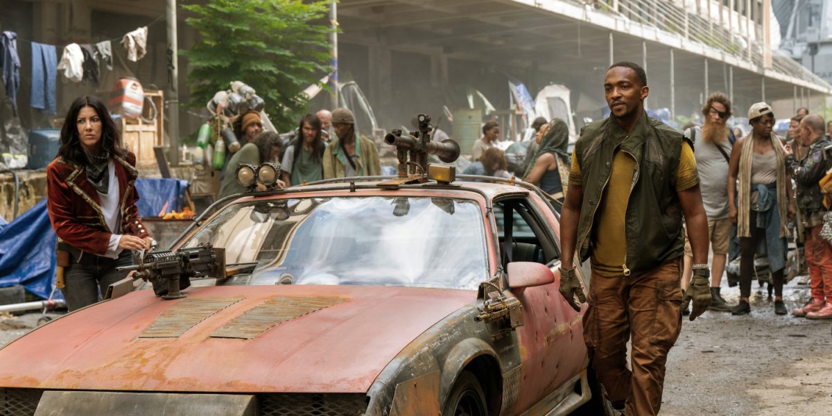 TWISTED-METAL-3 | film.org.pl Two survivors stand by a rusted, weapon-mounted car in a crowded post‑apocalyptic street.