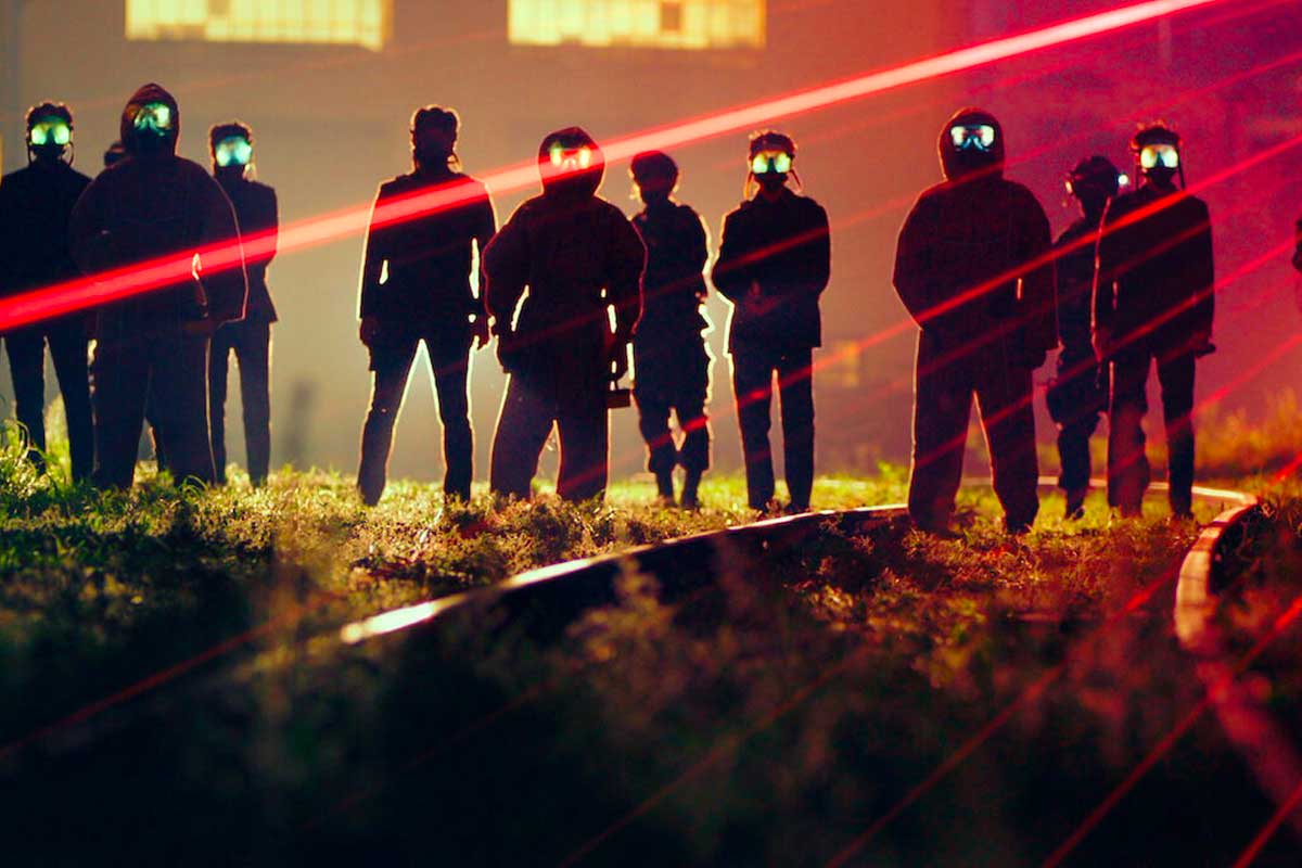 Silhouetted team in dark protective suits with glowing visors stands on a grassy area at night, red laser beams crossing the scene.