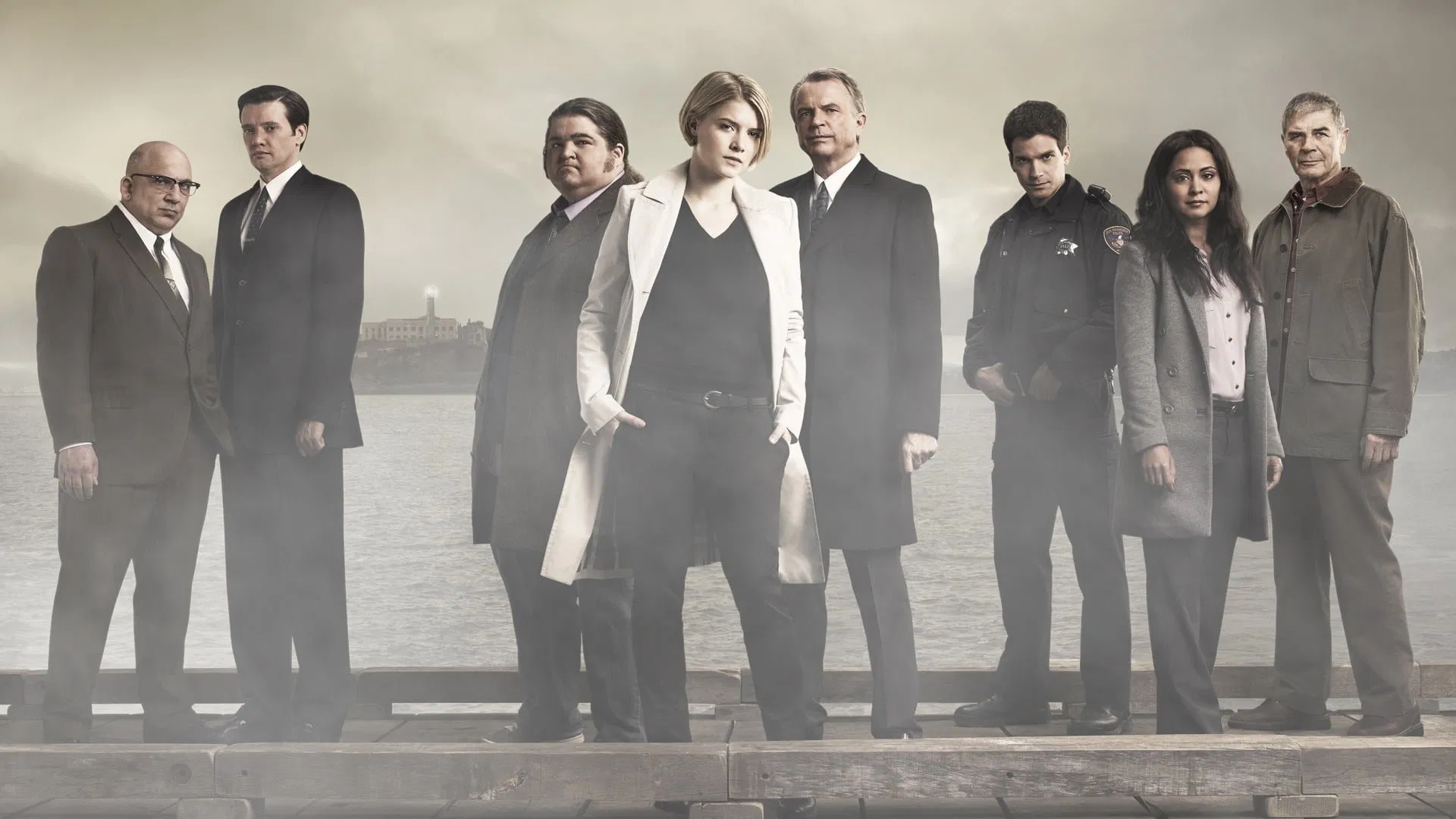 Group portrait of eight adults in business and law-enforcement attire standing on a wooden pier by a misty body of water, facing the camera with serious expressions.