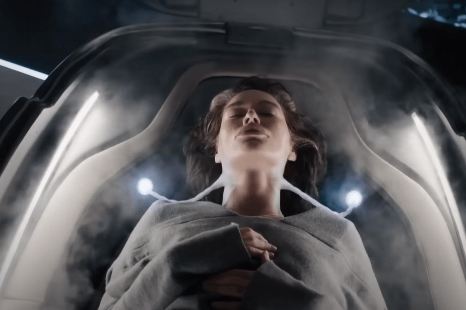 beacon-23-2 | film.org.pl Person lying inside a clear medical pod with eyes closed and hands folded over the chest amid mist.