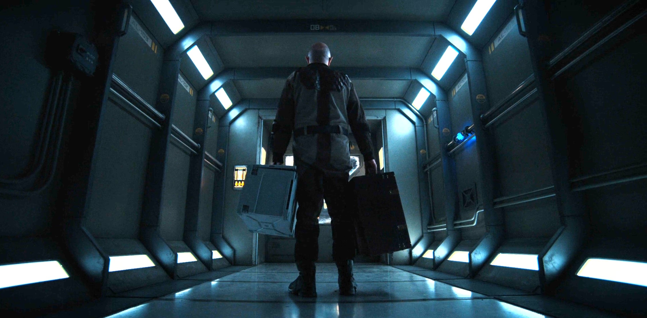 beacon-23-3 | film.org.pl Back view of a person in a gray and black suit walking down a blue-lit sci-fi corridor with two bags at their sides.