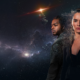 Two space explorers, a man and a woman, stand against a star-filled space backdrop; the woman appears to dissolve into particles on the right side.