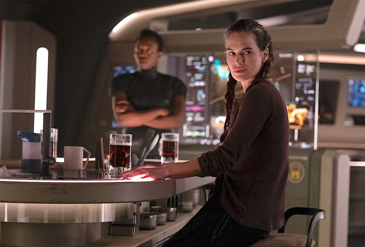 beacon-23 | film.org.pl Woman with braids sits at a sleek sci‑fi style bar, two red drinks on the counter, man in background.