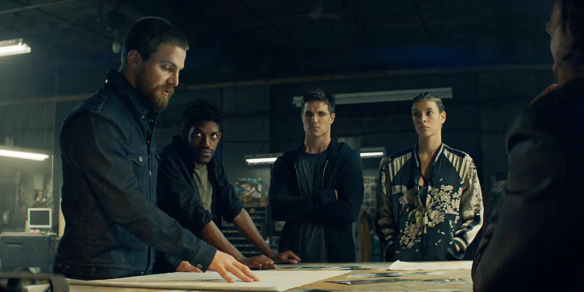 code-8-2 | film.org.pl A bearded man in a denim jacket explains plans on a large table while four teammates listen intently in a dim workshop.